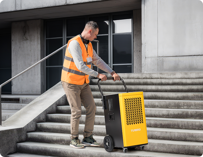 A worker in a safety vest is maneuvering a yellow dehumidifier down outdoor stairs.